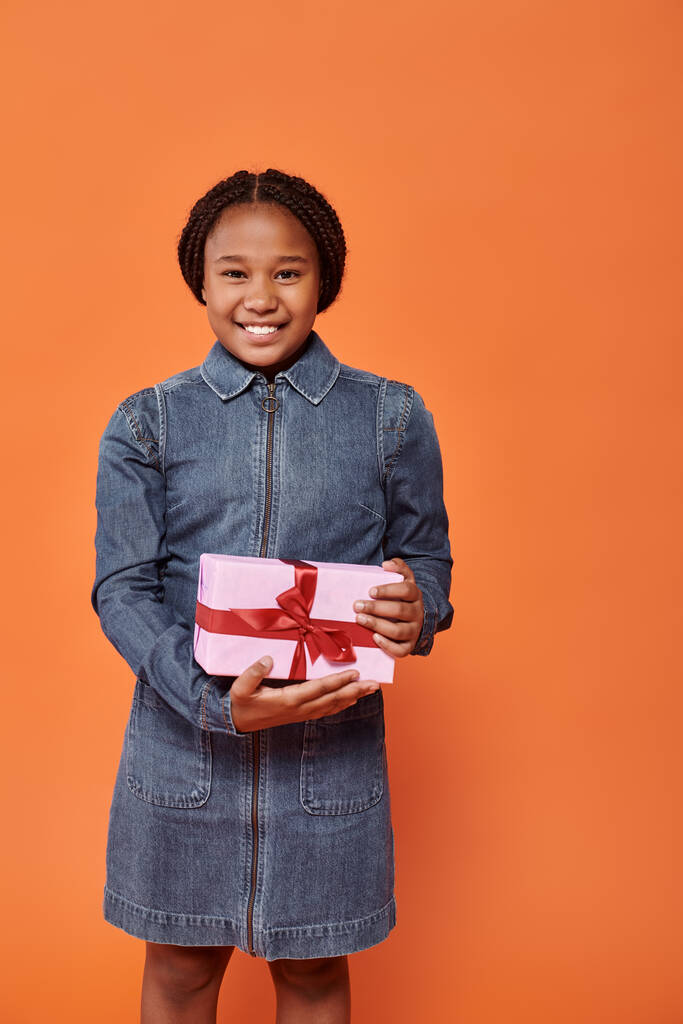 vrolijk Afrikaans amerikaans meisje in denim jurk holding cadeau en kijken naar camera op oranje - Foto, afbeelding
