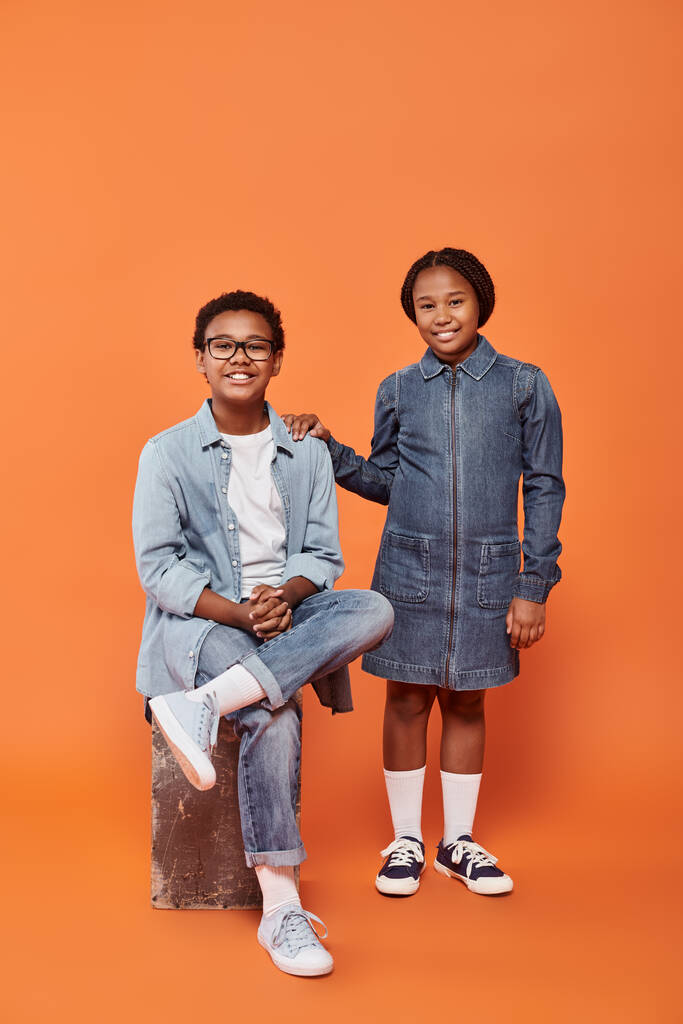 オレンジ色の背景で一緒にポーズするカジュアルなデニム服で陽気なアフリカ系アメリカ人の子供たち - 写真・画像
