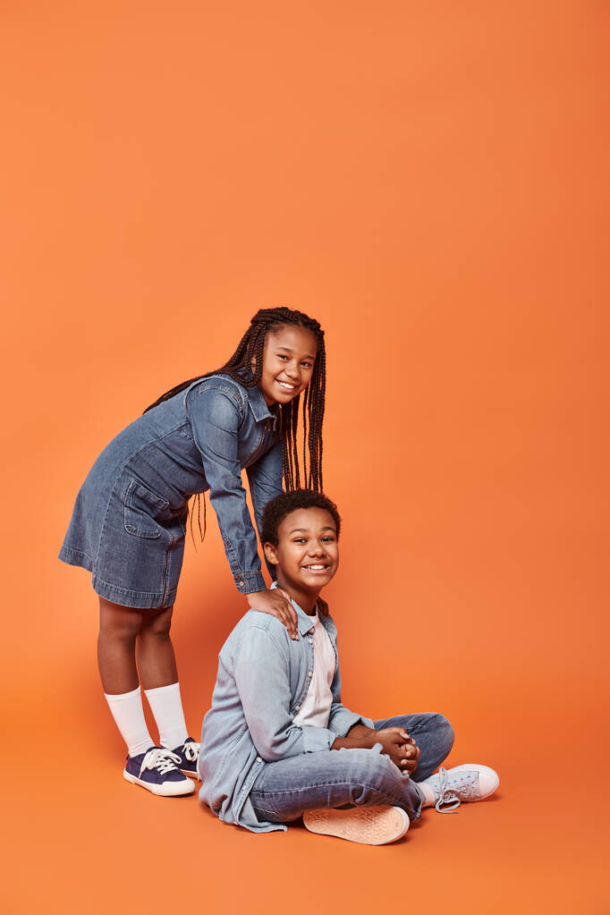 cheerful african american girl in casual denim attire standing and leaning on boy on orange - Photo, Image