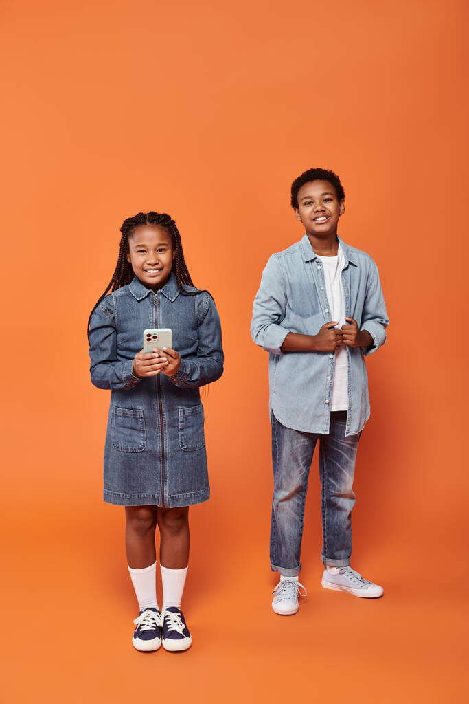 счастливая африканская американская девушка с косичками с помощью смартфона рядом веселый мальчик на оранжевом фоне - Фото, изображение