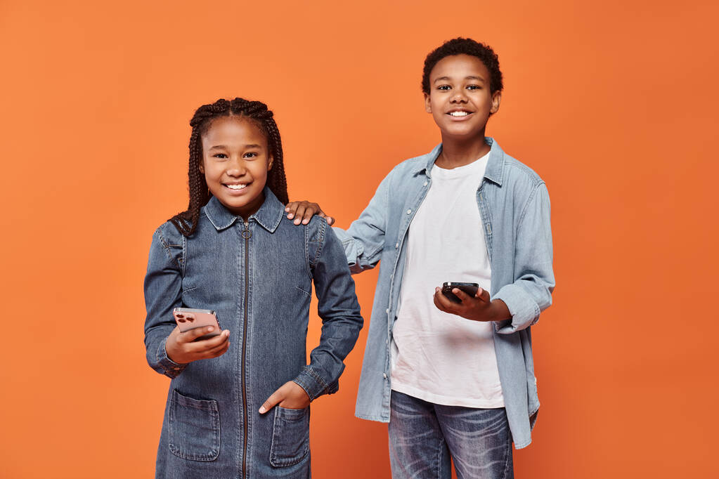Mutlu Afrikalı Amerikalı kız ve erkek günlük kıyafetleri içinde turuncu arka planda akıllı telefonlar taşıyorlar. - Fotoğraf, Görsel