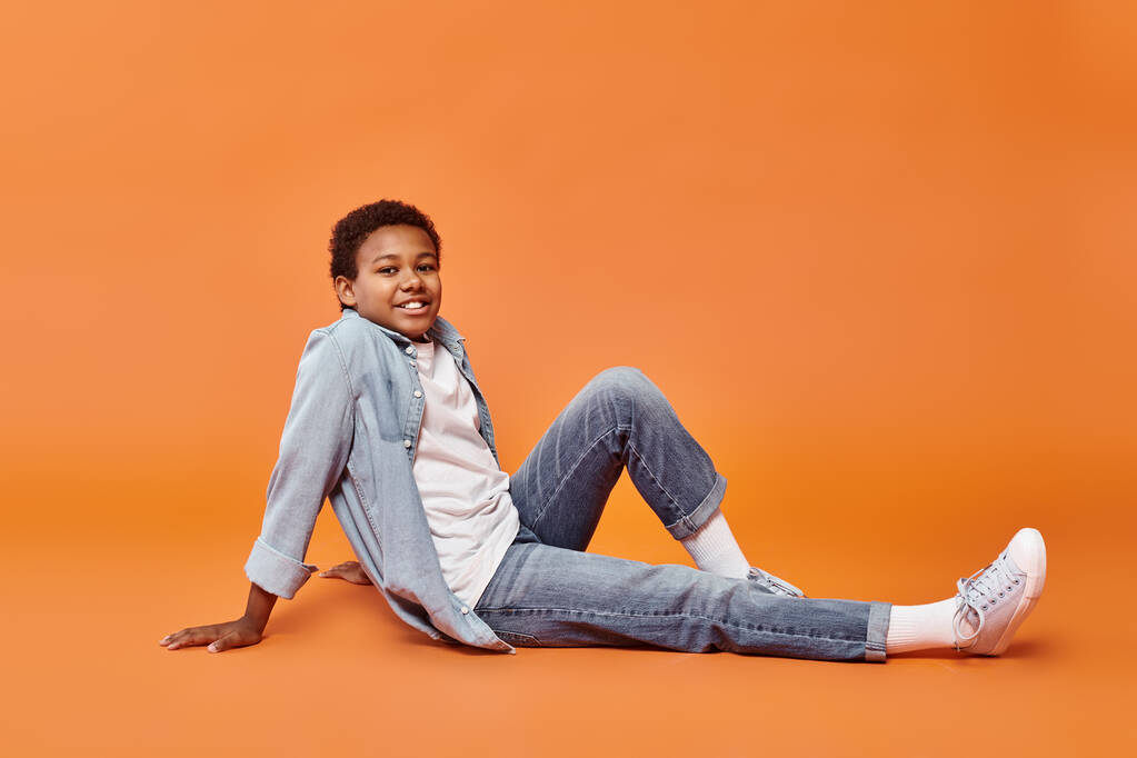 feliz menino americano africano pré-adolescente em attira casual sentado no chão em fundo laranja - Foto, Imagem