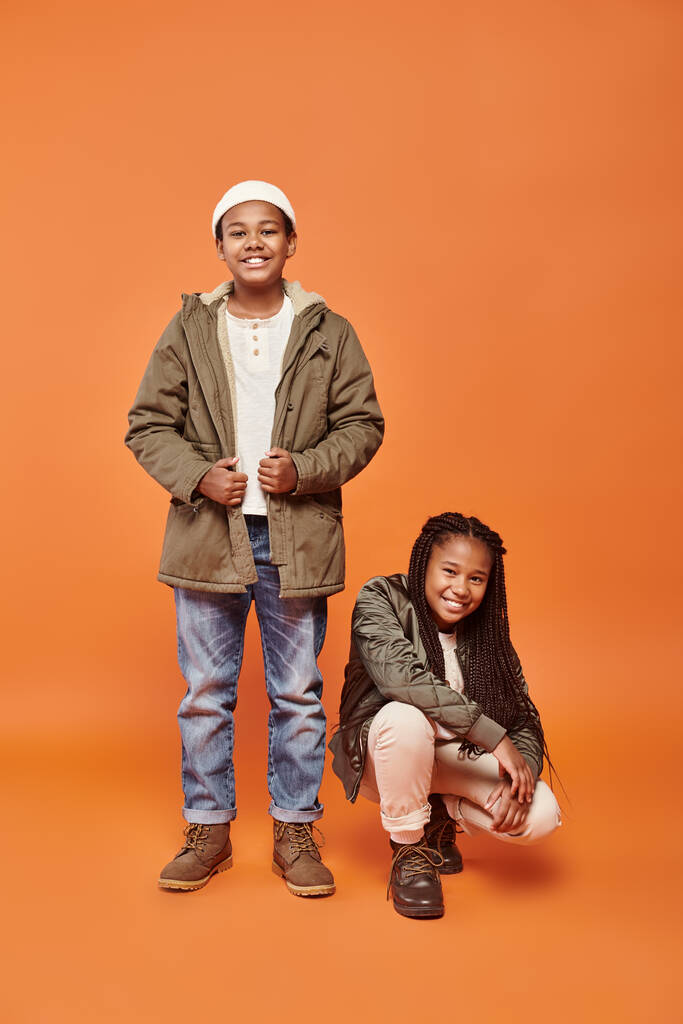 alegre pré-adolescente afro-americanos amigos em jaquetas quentes posando em movimento no fundo laranja - Foto, Imagem