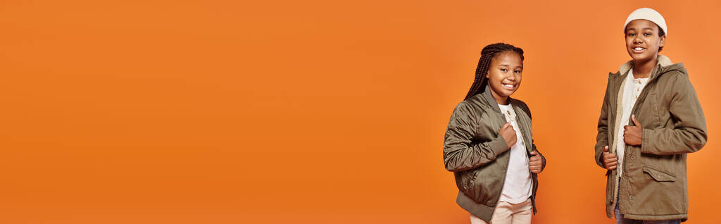 wesoły afrykański amerykański chłopiec i dziewczyna w ciepłych strojach uśmiecha się do kamery na pomarańczowym tle, baner - Zdjęcie, obraz