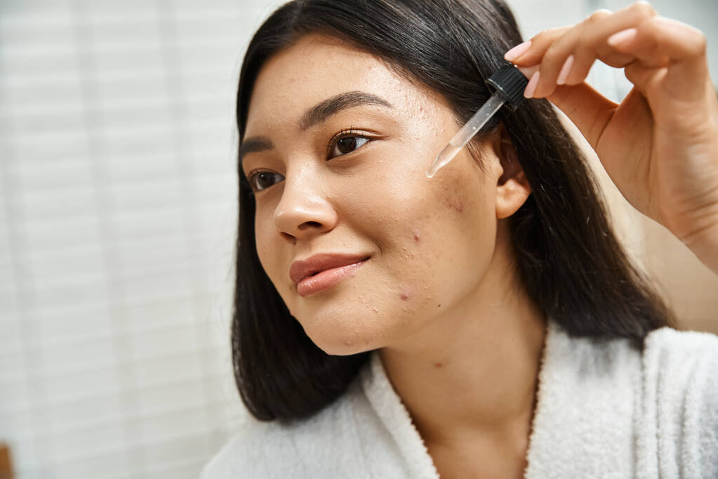 jovem e morena mulher asiática em roupão de banho aplicando soro facial para tratar manchas no rosto - Foto, Imagem