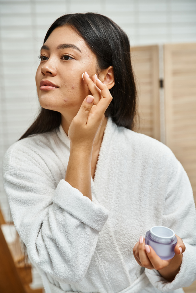 brunetky asijské žena s akné nanášení krém na obličej a odvrací pohled v koupelně, kožní problémy - Fotografie, Obrázek