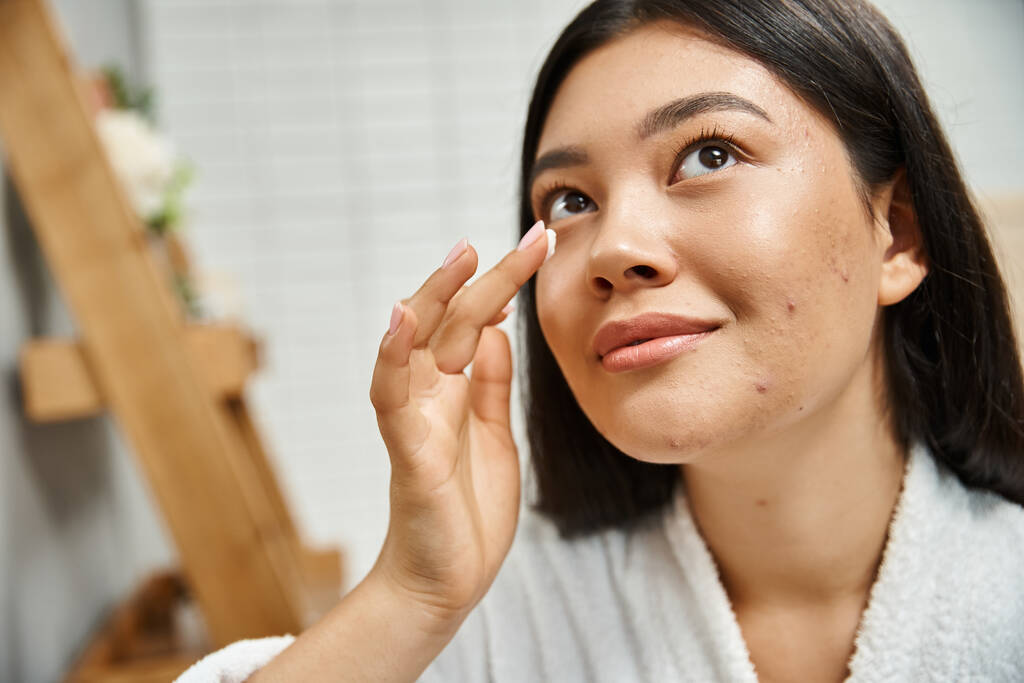 brünette asiatische Frau mit Akne Creme auf Gesicht auftragen und sich im Badezimmer, Hautprobleme - Foto, Bild
