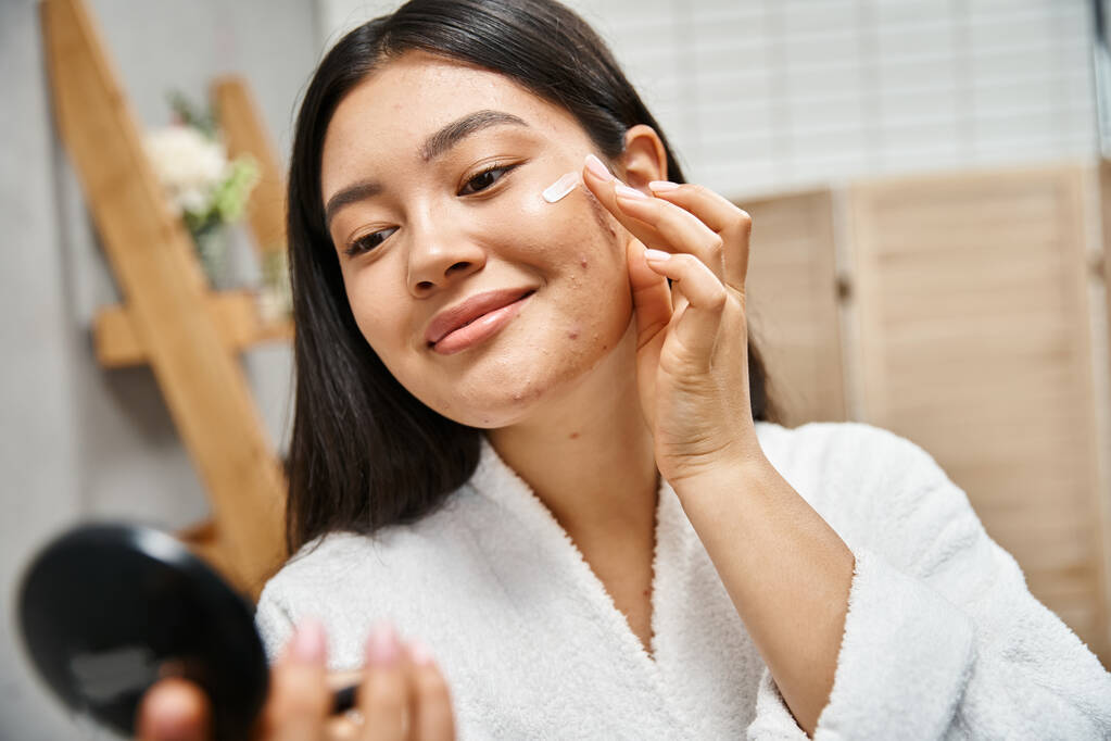 jovem mulher asiática com cabelo morena aplicando creme de tratamento de acne e olhando para espelho compacto - Foto, Imagem