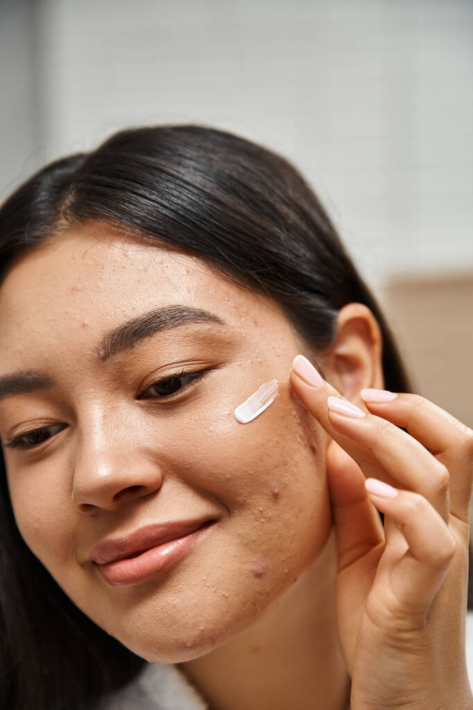 foto ravvicinata di giovane donna asiatica con capelli castani che applica crema per il trattamento dell'acne sul viso - Foto, immagini