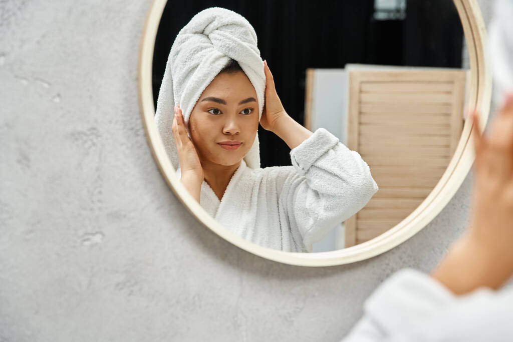 молодая азиатская женщина с полотенцем на голове и акне склонной кожи, глядя на зеркало ванной в доме - Фото, изображение