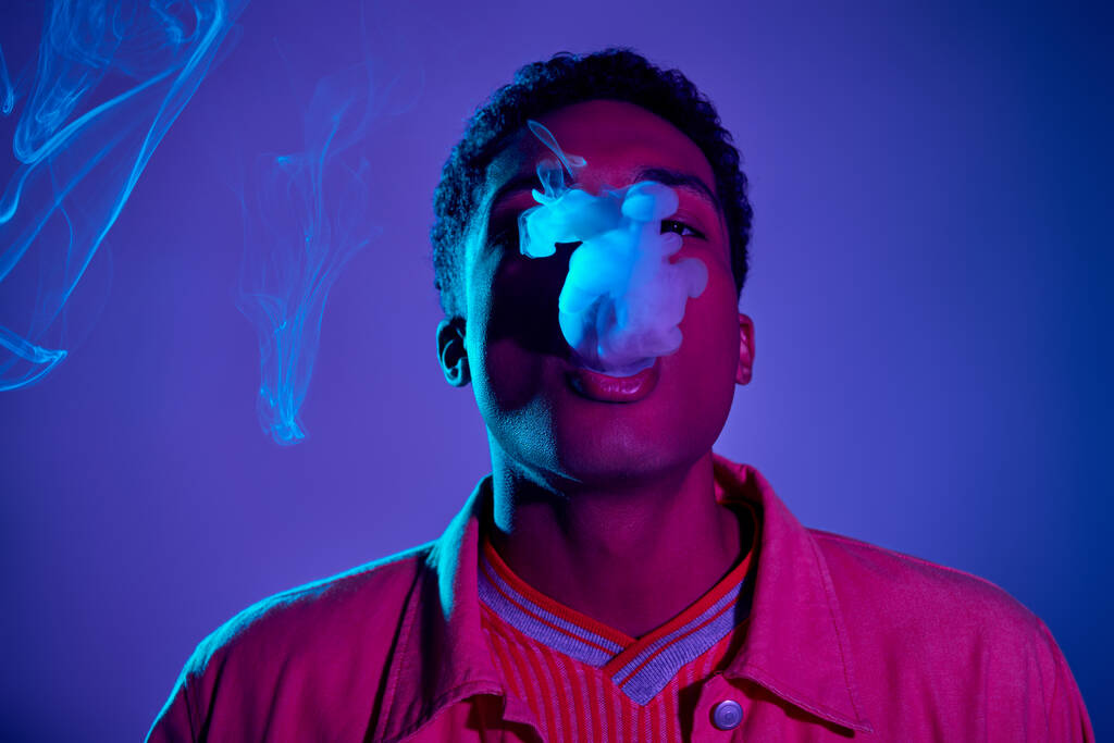 tipo afroamericano con estilo exhalando humo contra fondo azul con iluminación púrpura, gen z - Foto, Imagen