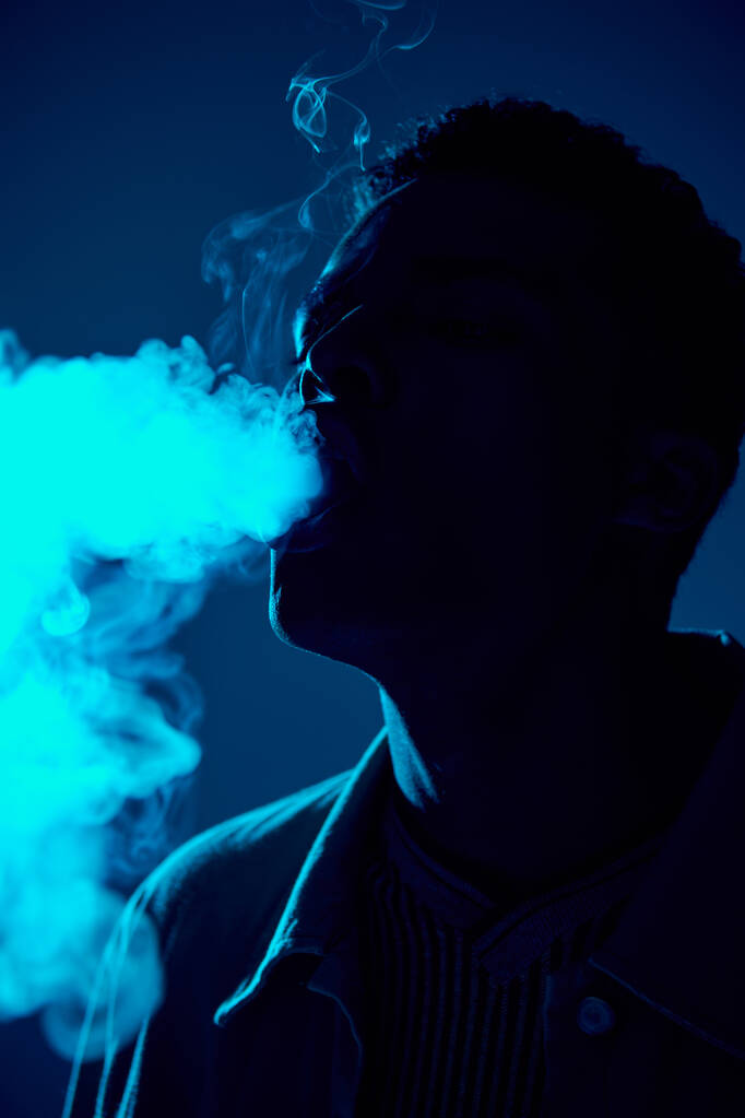 retrato del hombre afroamericano exhalando humo contra fondo oscuro con luz azul, vapeo - Foto, imagen