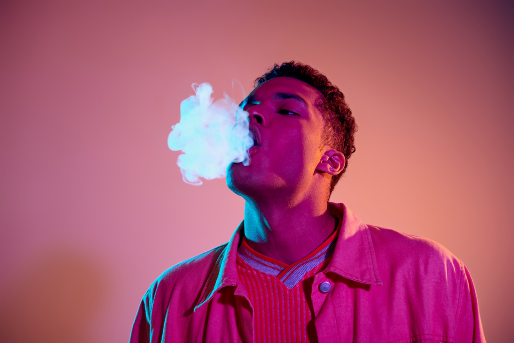 retrato del hombre afroamericano exhalando humo contra un fondo vibrante con iluminación azul, vapor - Foto, imagen