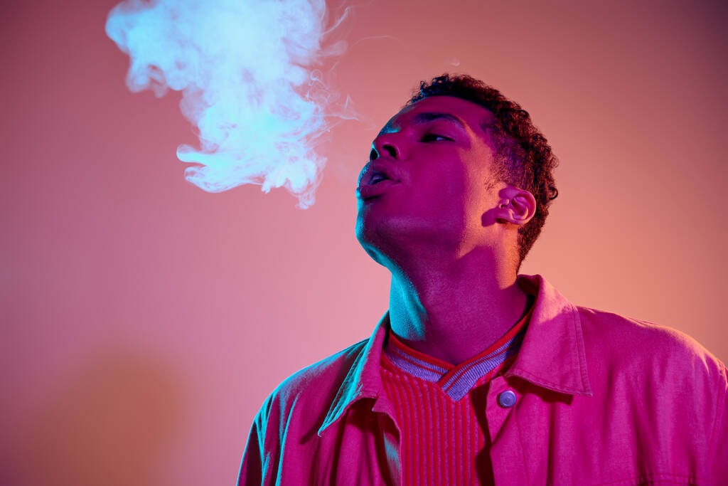 ritratto dell'uomo afroamericano che esala fumo su uno sfondo vibrante con illuminazione blu, vaporizzatore - Foto, immagini