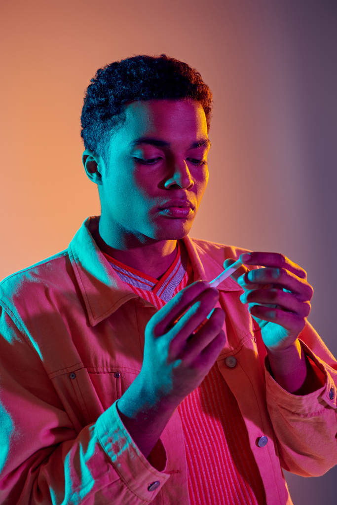 Afrikanisch-amerikanischer Kerl schaut auf Zigarettenrolle auf buntem Hintergrund mit blauer Neonbeleuchtung - Foto, Bild