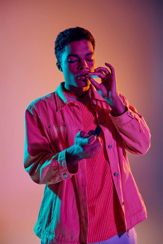 uomo afroamericano accendendo una sigaretta mentre tiene accendino sotto luci al neon blu su rosa - Foto, immagini