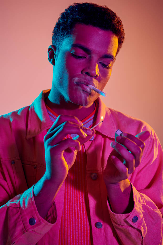 ピンクの背景に青いネオンライトの下でマッチとタバコを照らすアフリカ系アメリカ人男性 - 写真・画像