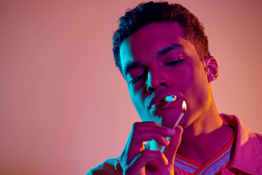 africano americano chico iluminación un cigarrillo con partido bajo azul neón luces sobre rosa fondo - Foto, Imagen