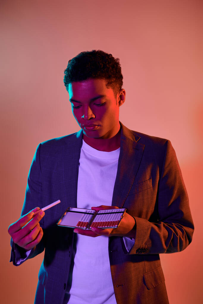 青い照明でピンクの背景にタバコのケースを保持している若いアフリカ系アメリカ人の肖像画 - 写真・画像
