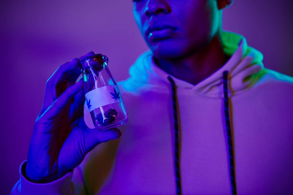hombre afroamericano en sudadera con capucha sosteniendo tarro con cannabis medicinal sobre fondo azul oscuro - Foto, Imagen