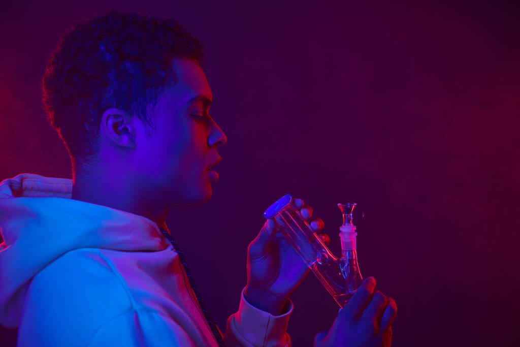 young african american man in hoodie looking at glass bong on dark purple background with lighting - Photo, Image