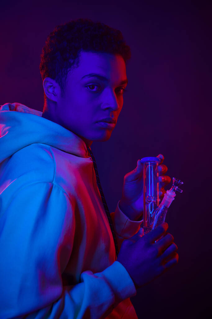молодий афроамериканський чоловік в светрі дивиться на скляний бонг на темно-синьому фоні з освітленням - Фото, зображення