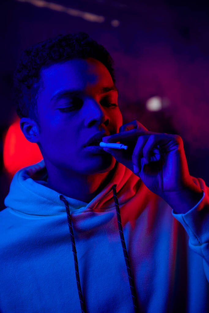 アフリカ系アメリカ人男性が真っ暗な青い背景に赤い光で口の近くで巻いたタバコを抱えています - 写真・画像