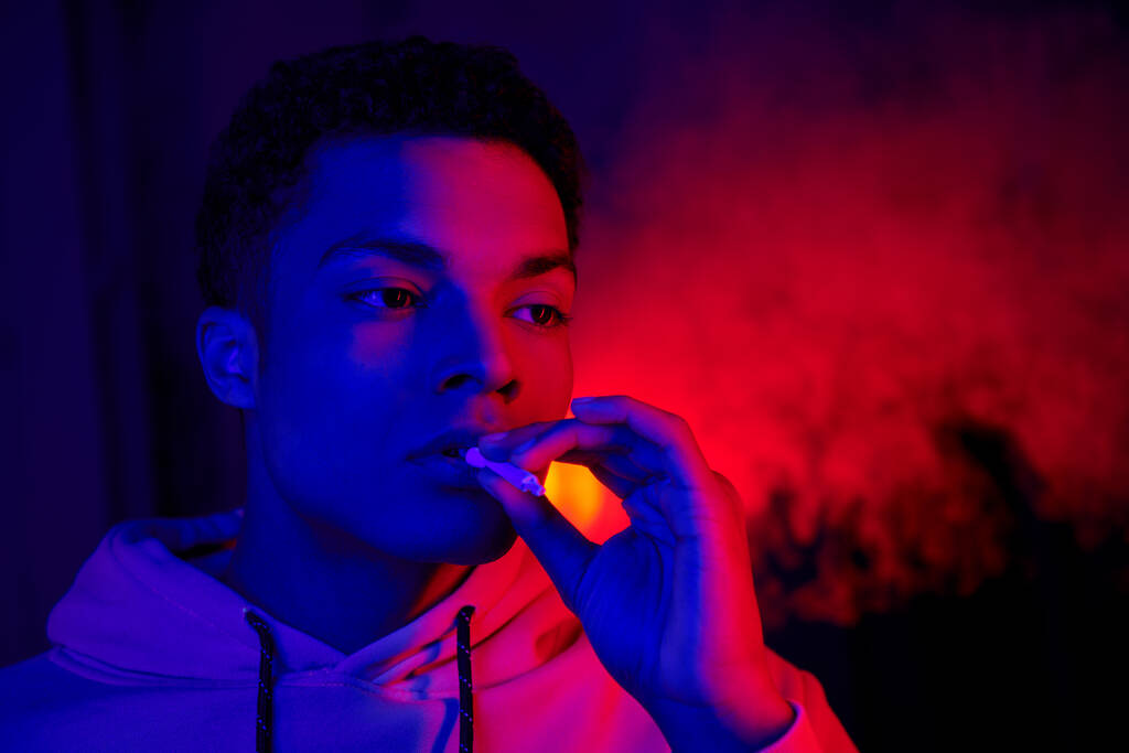 viileä afrikkalainen amerikkalainen mies pitämällä rullattu savuke lähellä suuta tummansininen punainen valo, tupakoitsija - Valokuva, kuva