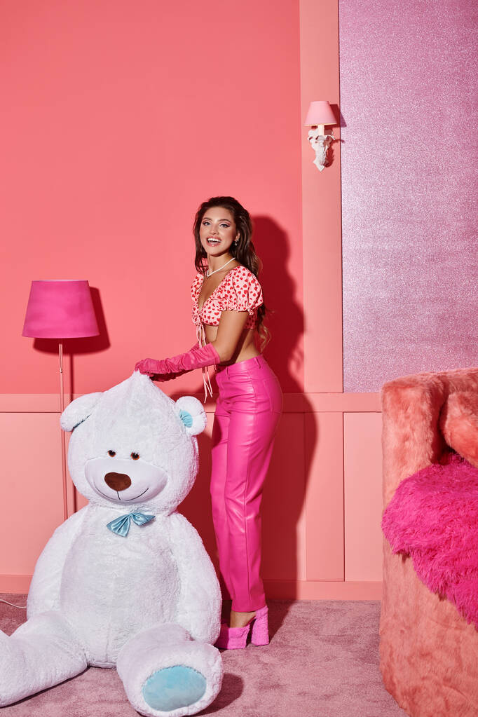 fröhliche junge Frau in rosa bauchfreiem Top und Hose lacht in der Nähe eines riesigen Teddybären in einem lebhaften Raum - Foto, Bild