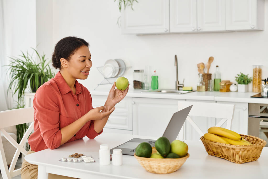 幸せなアフリカのアメリカの栄養士は,キッチンでラップトップにダイエットアドバイスをしながらリンゴを保持しています - 写真・画像