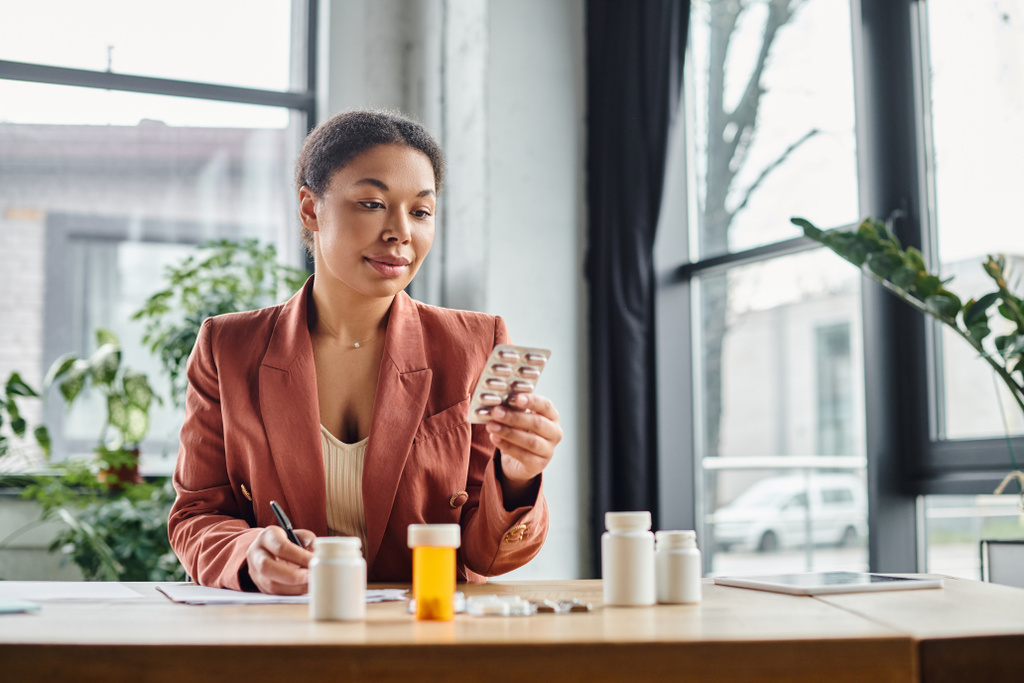 krullend afrikaanse Amerikaanse voedingsdeskundige evalueren medicatie tijdens het schrijven van notities in haar kantoor - Foto, afbeelding