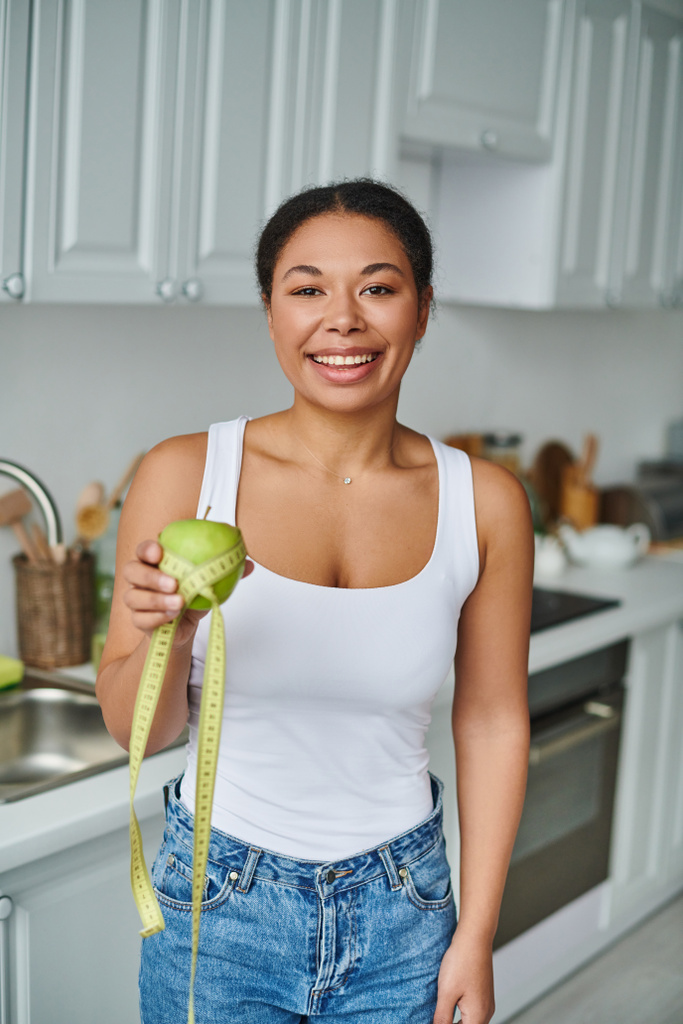 mujer afroamericana feliz con cinta métrica y manzana promoviendo una dieta saludable en la cocina - Foto, imagen