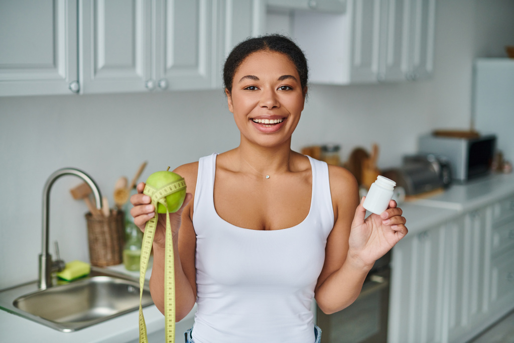 happy african american woman with measuring tape with apple and bottle with supplements - Photo, Image
