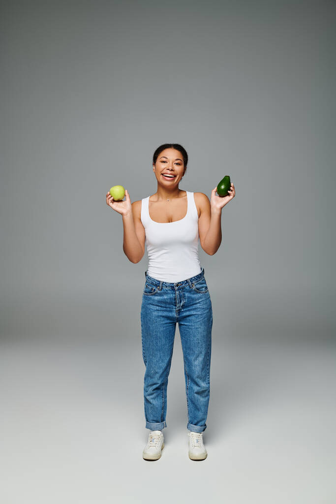 gelukkig Afrikaans amerikaanse vrouw met appel en avocado bevordering van evenwichtige voeding op grijze achtergrond - Foto, afbeelding