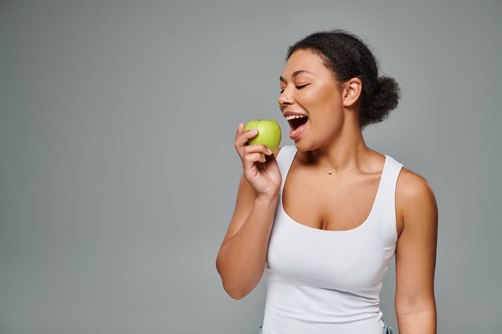 白い歯を持つ幸せなアフリカ系アメリカ人女性は緑のリンゴを灰色の背景で噛み,健康的な食事 - 写真・画像