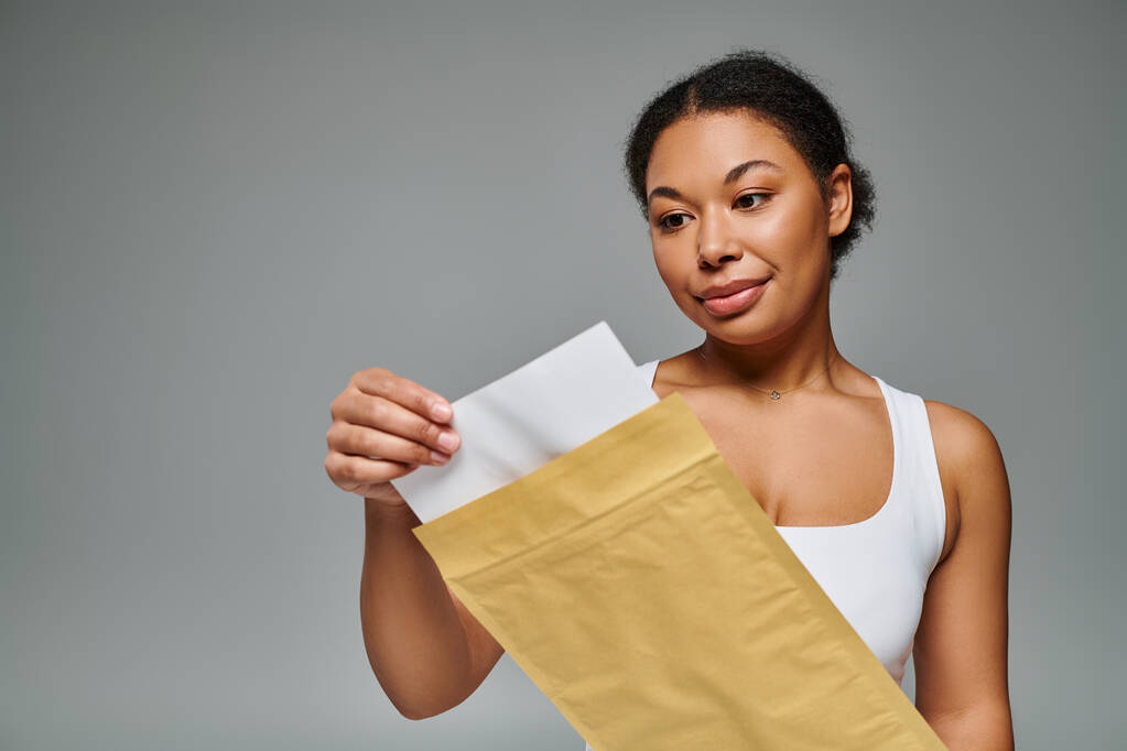 fröhlich afrikanisch-amerikanische Diätassistentin hält Umschlag mit Ernährungsplan vor grauem Hintergrund - Foto, Bild