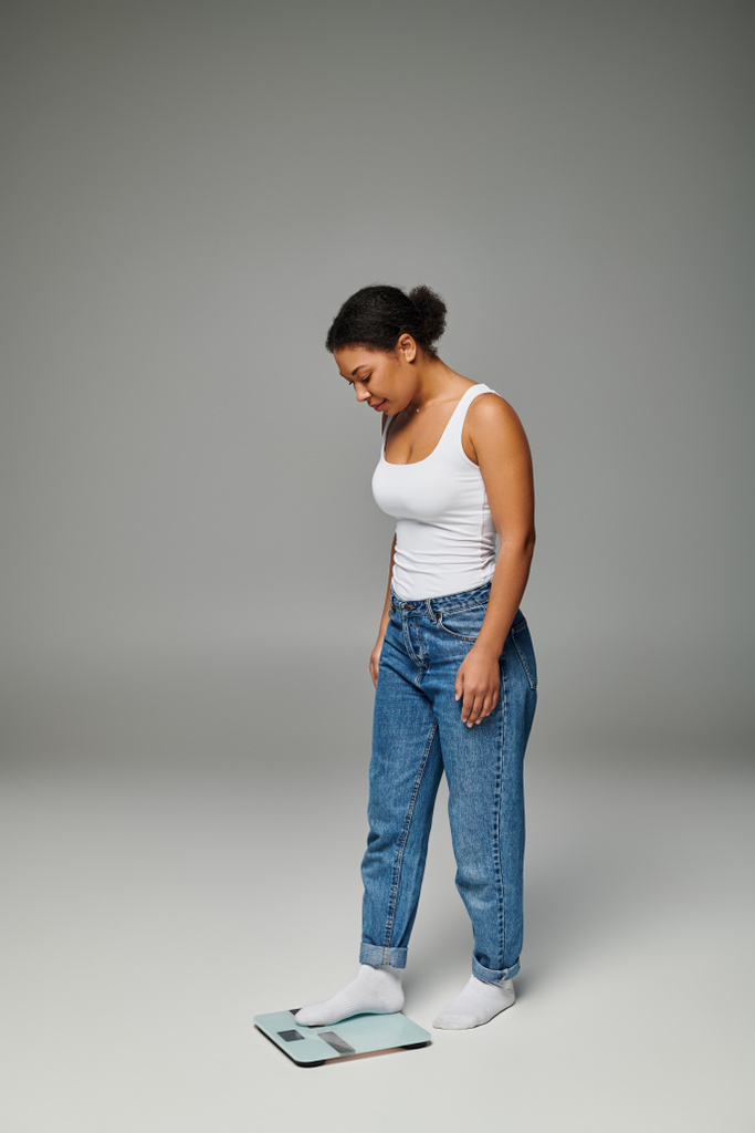 happy and young african american woman in casual attire standing on scales, grey background - Photo, Image