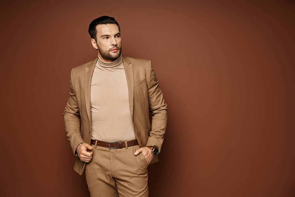 hombre guapo en traje elegante mirando hacia otro lado mientras posando con la mano en el bolsillo sobre fondo beige - Foto, imagen