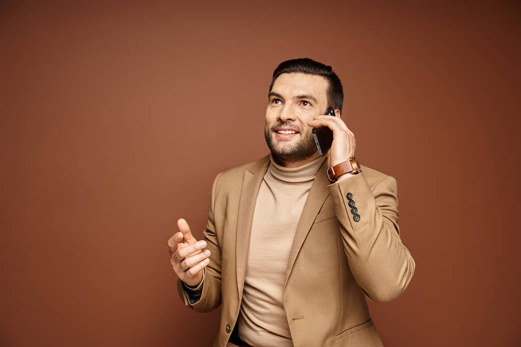 Stylish and handsome man in blazer smiling and having phone call on smartphone, beige backdrop - Photo, Image