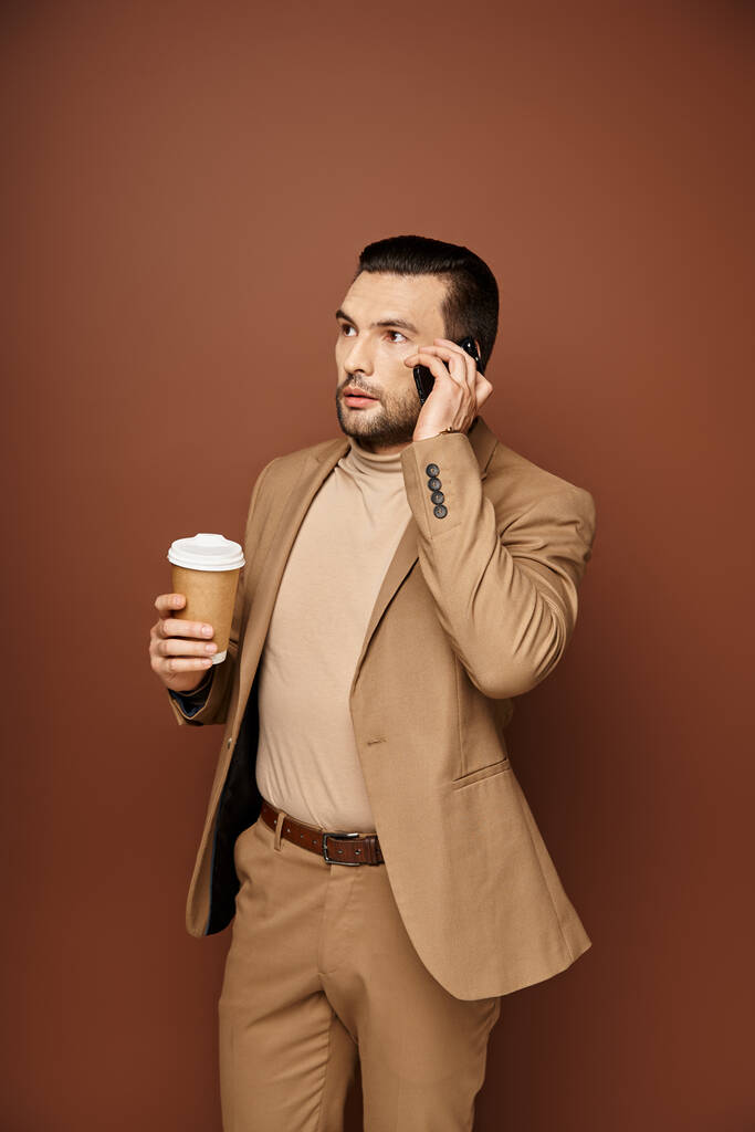 hombre guapo en traje elegante celebración de café para ir y tener llamada en el teléfono inteligente en fondo beige - Foto, Imagen