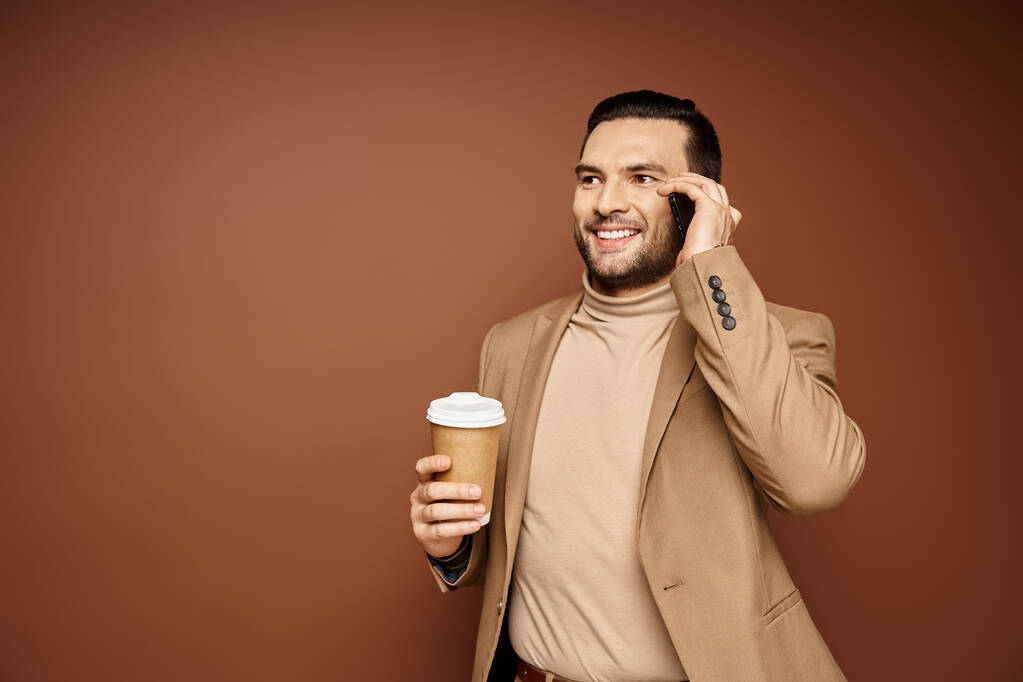 gelukkig man in stijlvolle kleding houden van koffie te gaan en het hebben van oproep op smartphone op beige achtergrond - Foto, afbeelding