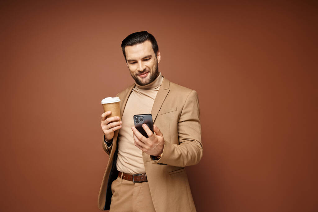 ευτυχισμένος άνθρωπος με κομψή ενδυμασία κρατώντας καφέ για να πάει και να χρησιμοποιήσει το smartphone του σε μπεζ φόντο - Φωτογραφία, εικόνα