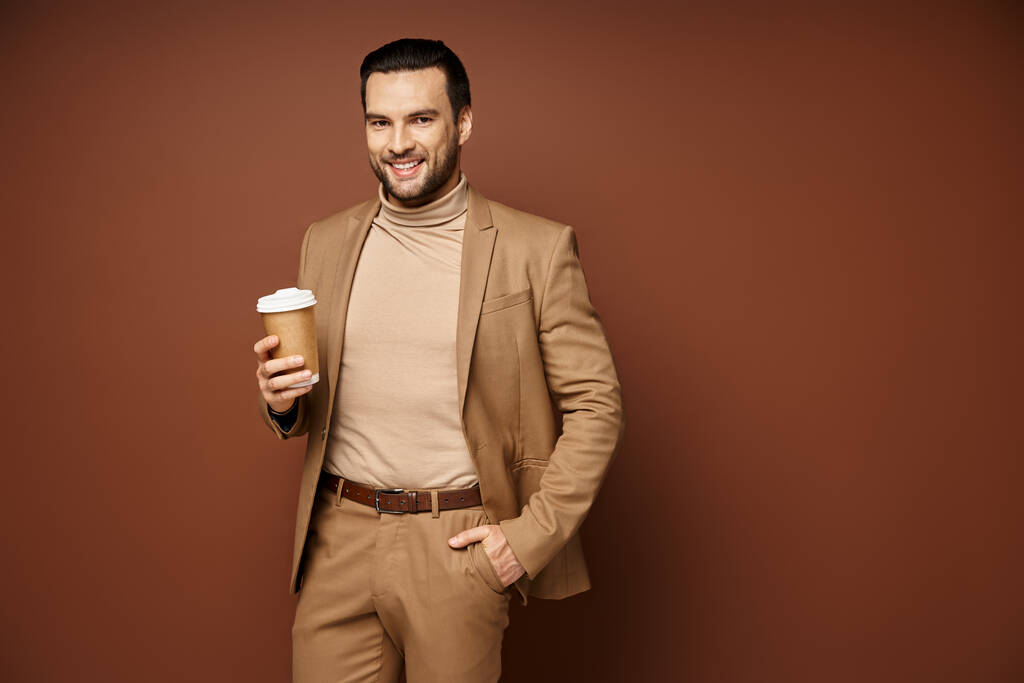 ευτυχισμένος άνθρωπος σε κομψή ενδυμασία κρατώντας καφέ για να πάει και στέκεται με το χέρι στην τσέπη σε μπεζ φόντο - Φωτογραφία, εικόνα
