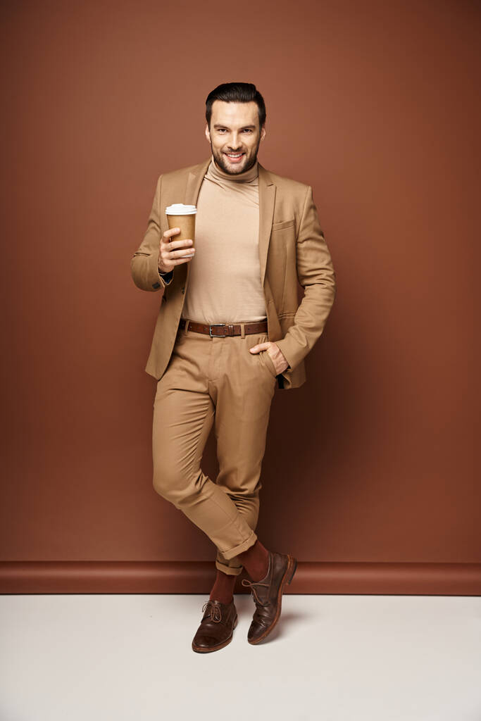 contenuto uomo in abbigliamento elegante in possesso di caffè per andare e in posa con mano in tasca su sfondo beige - Foto, immagini