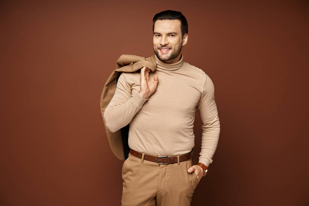 hombre feliz con cuello alto sosteniendo su chaqueta sobre el hombro y posando con la mano en el bolsillo en beige - Foto, imagen