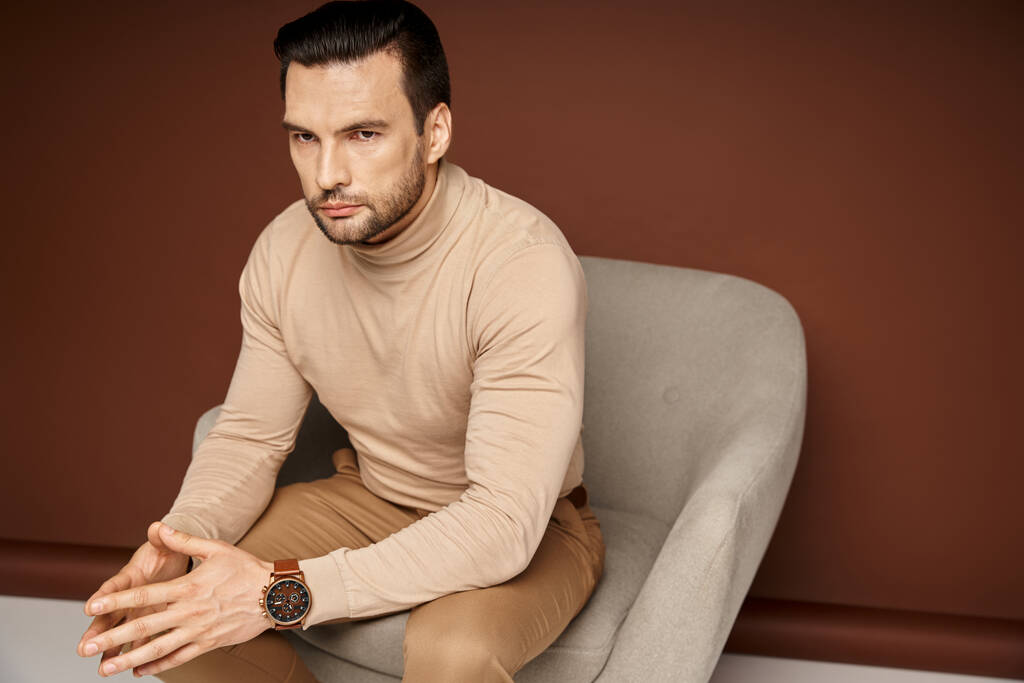 handsome and serious man in turtleneck sitting on comfortable armchair on beige background - Photo, Image