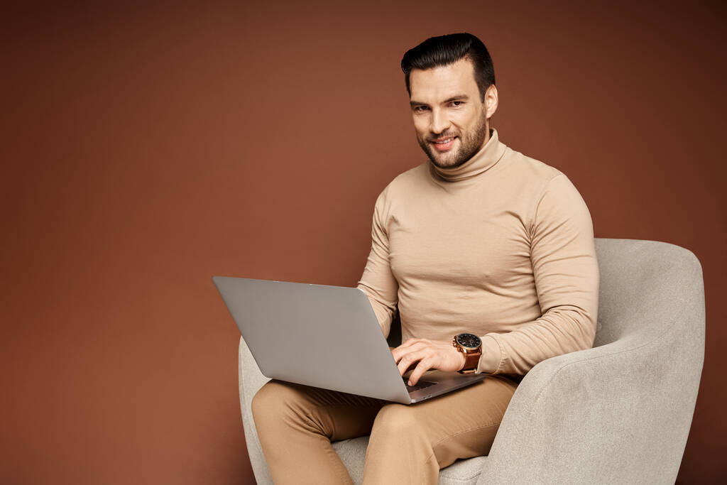 happy man in turtleneck sitting on comfortable armchair and using laptop on beige backdrop - Photo, Image