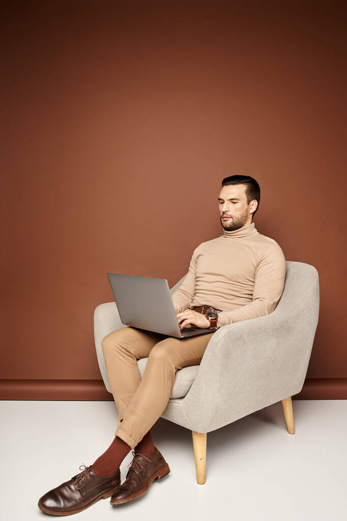gut aussehender Mann im Rollkragen sitzt auf bequemen Sesseln und benutzt Laptop auf beigem Hintergrund - Foto, Bild