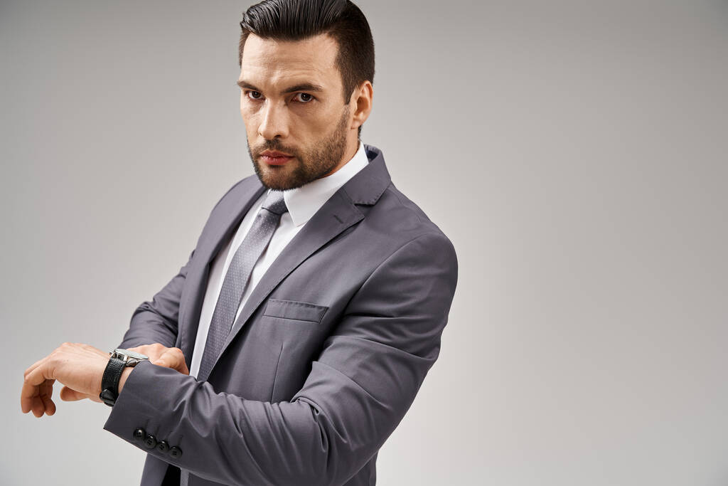 hombre de negocios guapo posando en traje elegante revisando su reloj de pulsera sobre fondo gris, carrera - Foto, imagen