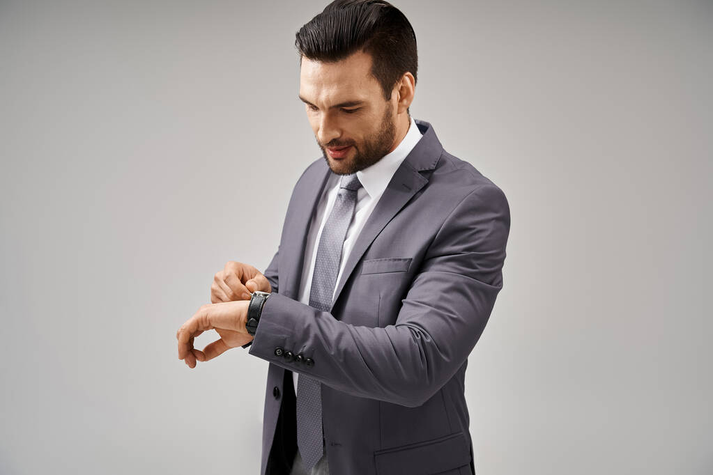 красивый бизнесмен позирует в элегантном костюме, проверяя свои наручные часы на сером фоне, пунктуальный - Фото, изображение